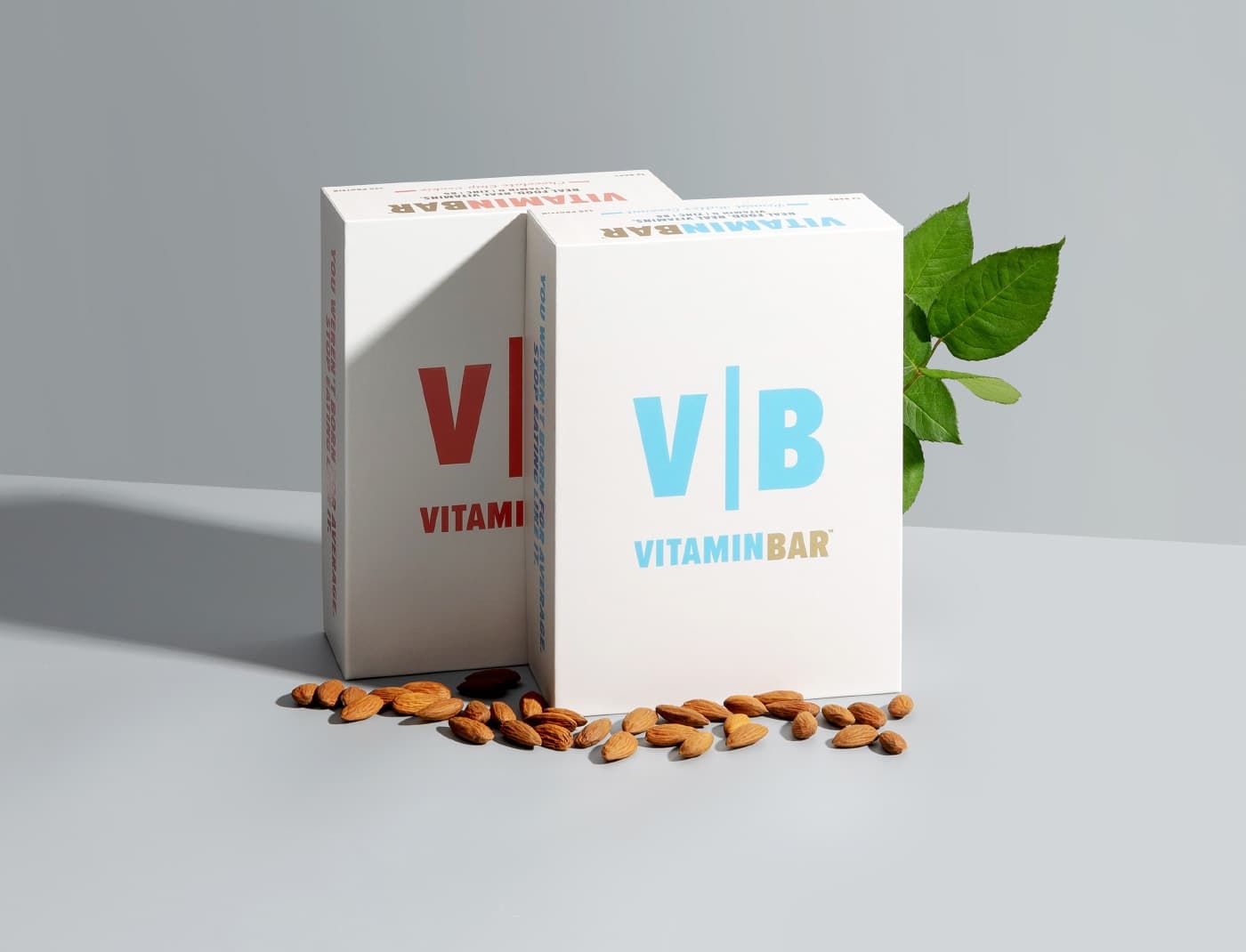 Two upright VitaminBar boxes in red and blue standing on a grey surface with scattered almonds and green leaves.