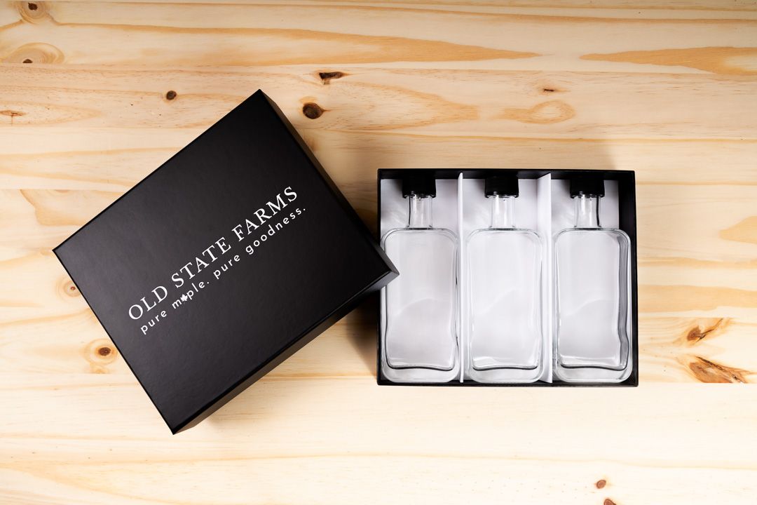 Top-down view of an open Old State Farms rigid box containing three empty glass syrup bottles separated by white dividers.