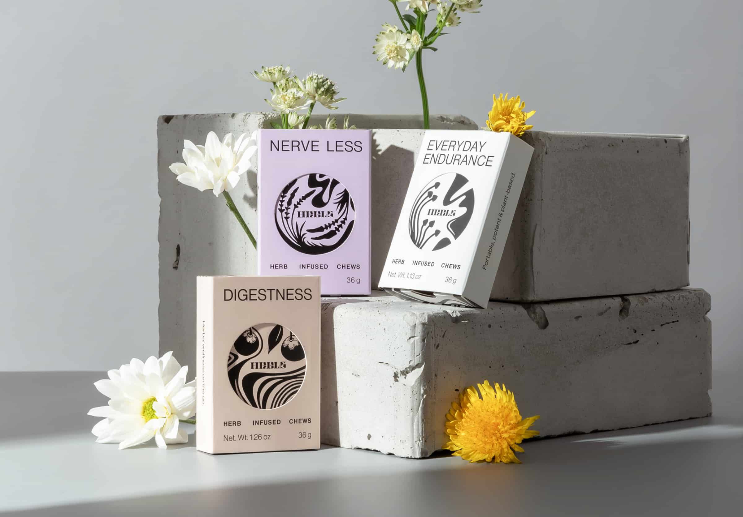 Three HRBLS herbal chew boxes in purple, white, and beige arranged on concrete blocks with white flowers.