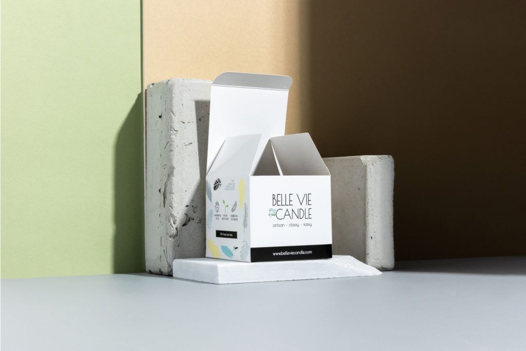 Open white Belle Vie Candle folding carton displaying side icons, resting on concrete blocks against a color-blocked wall.