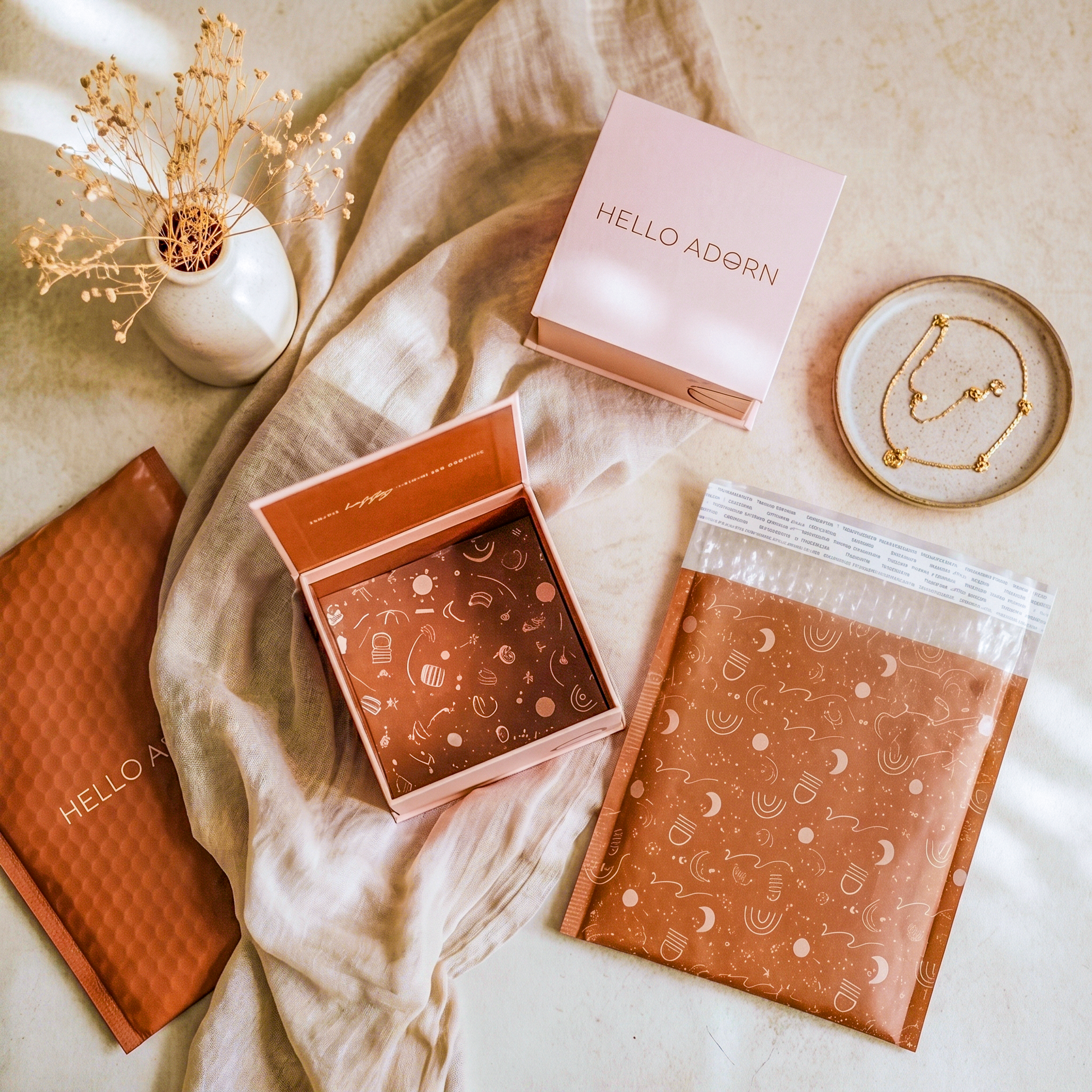 A flat lay of Hello Adorn jewelry packaging on a draped beige cloth. It features two light pink boxes, rust-colored patterned bubble mailers, a delicate gold necklace in a small ceramic dish, and dried flowers in a white vase.