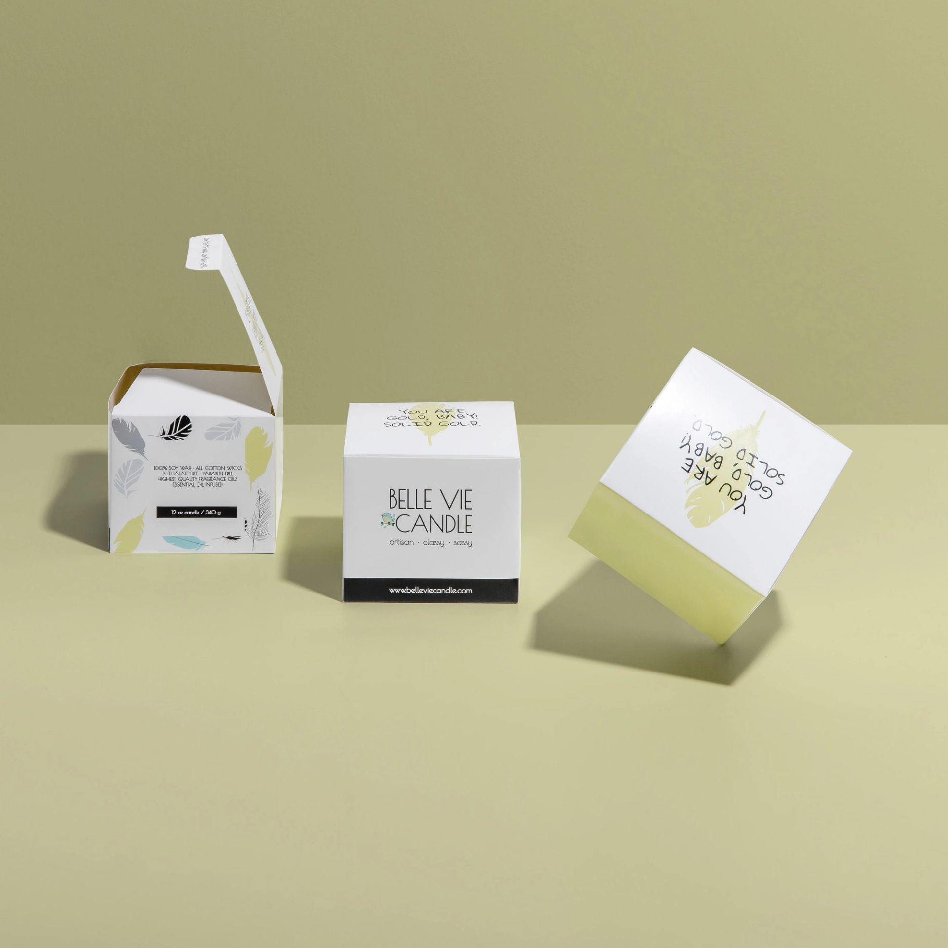 Three Belle Vie Candle boxes on a green surface; one open, one closed, and one tumbling to reveal a printed top flap.