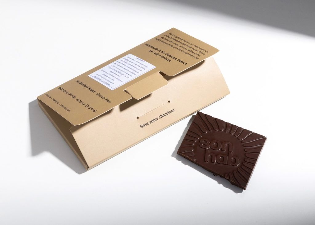 An open Sonhab chocolate wrapper laying flat next to a molded chocolate bar featuring a sunburst logo design.