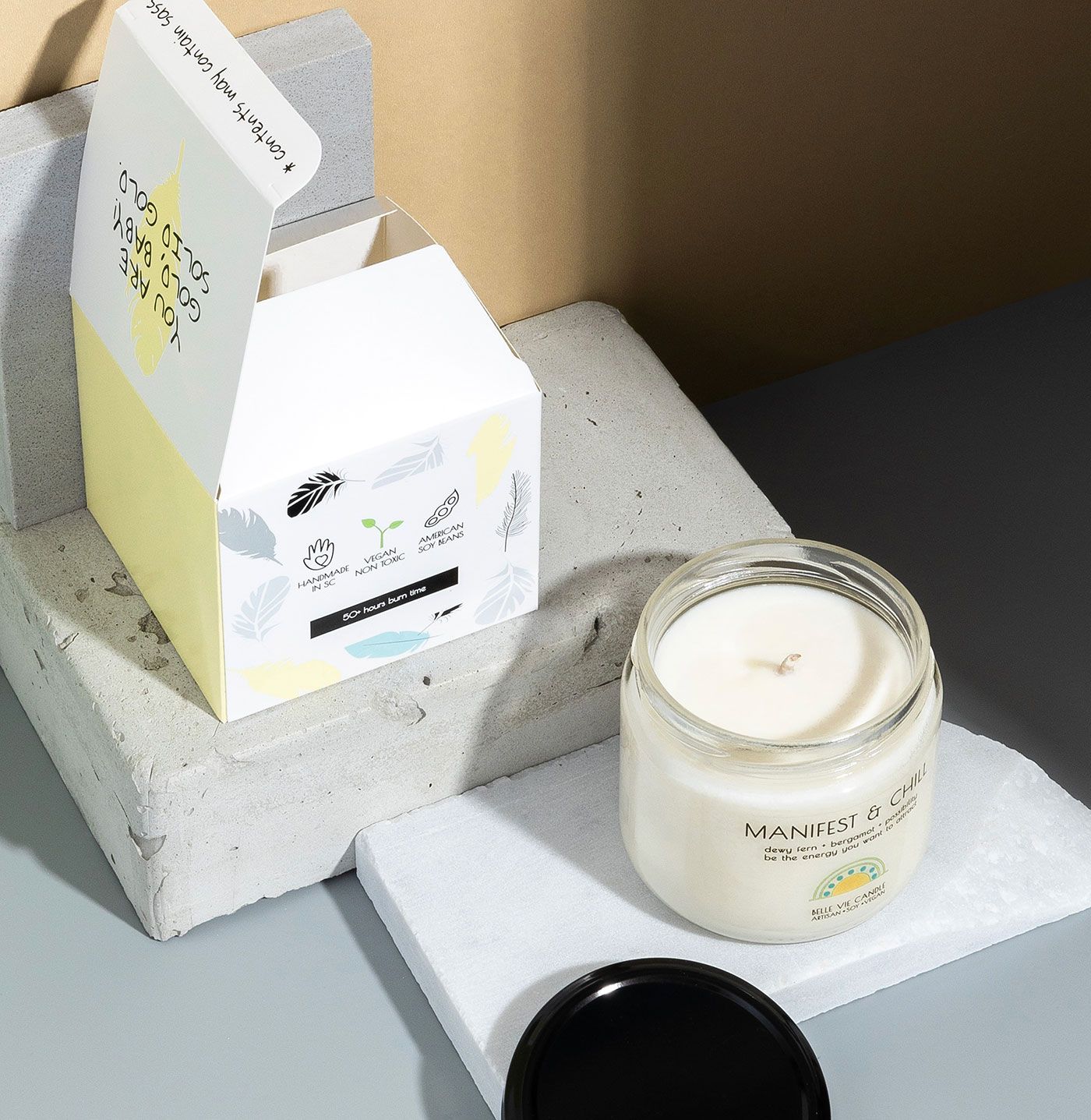 Angled view of a lit Belle Vie candle and its open packaging, showing a yellow interior flap with a compliment.