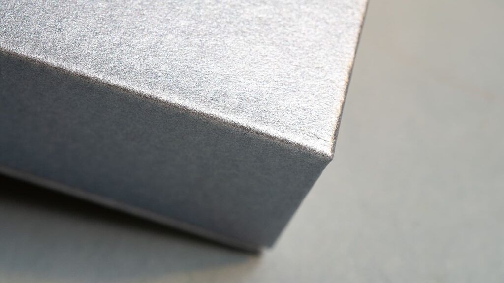 Macro close-up photograph showing the sharp, precision-folded corner of a custom presentation box made from textured metallic silver paper.
